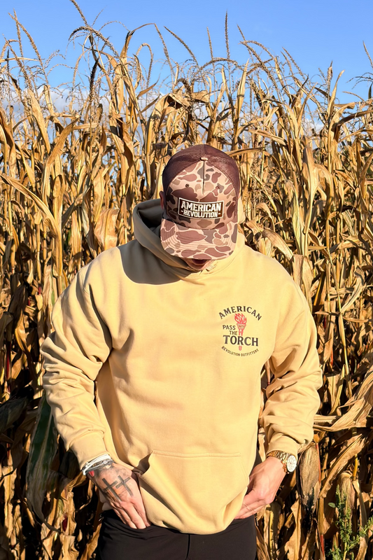 Person wearing a beige hoodie with 'American Torch' logo walking through a cornfield.