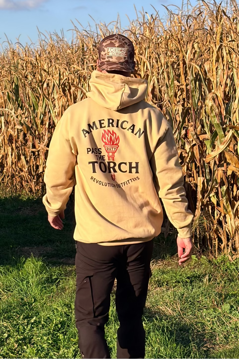 Person wearing a beige hoodie with 'American Torch' logo walking through a cornfield.