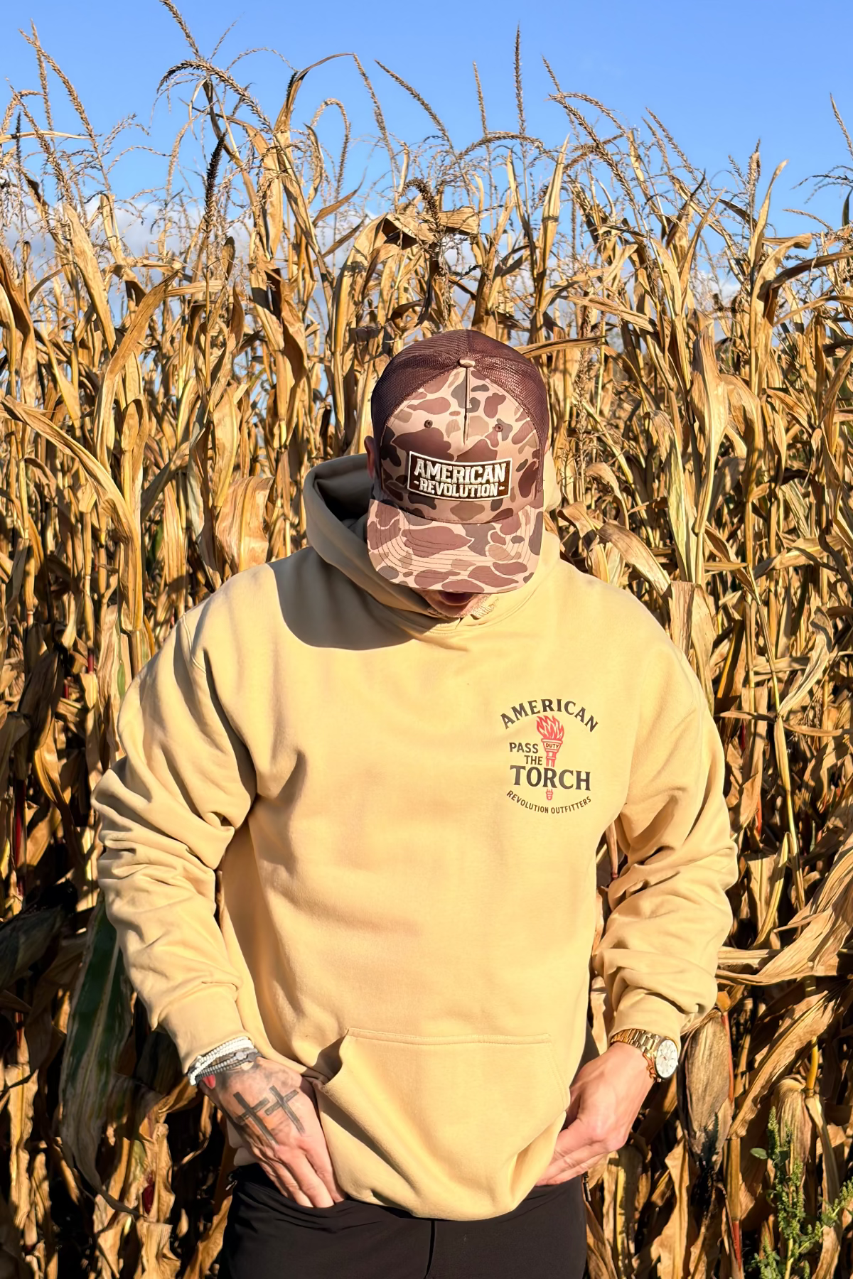 Person wearing a beige hoodie with 'American Torch' logo walking through a cornfield.